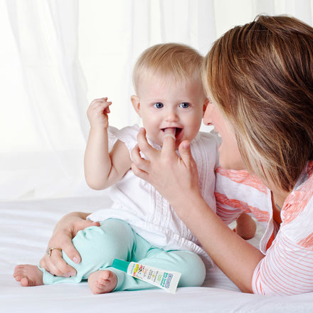 A woman gently applies Dr. Talbot's Baby Tooth & Gum Cleaner—made with natural, fluoride-free ingredients—to a smiling baby’s mouth as they sit on a bed, illuminated by natural light through white curtains.