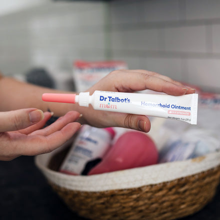 A hand holds a tube of Dr. Talbot’s Mom Pregnancy & Postpartum Hemorrhoid Ointment with an applicator tip attached, over a basket of health supplies.