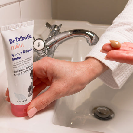 A person in a white robe holds Dr. Talbot’s Mom Vegan Nipple Balm near a sink labeled "hot," with a dab on their fingertip. The Ultimate Breastfeeding Care Package by Dr. Talbot's Mom is nearby, creating a nurturing scene.