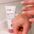 A hand with light pink nails applies a clear balm to another hand. In the background, Dr. Talbot's Vegan Nipple Balm, a gentle lanolin-free balm for breastfeeding moms, is visible on the bathroom counter.