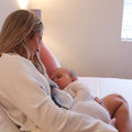 A woman with long blonde hair in a light robe sits on a bed, breastfeeding her baby in white pajamas. She uses Dr. Talbot's Silicone Breast Pump hands-free—a must-have for new moms—in a calm, softly lit room.