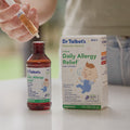 A hand holds a filled oral syringe above an open bottle of Dr. Talbot’s Infant Daily Allergy Relief, positioned next to the product box.