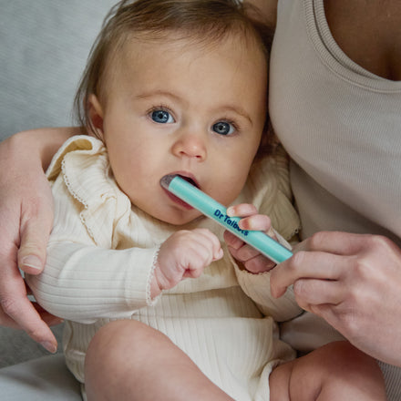An adult holds a baby who is chewing on a light blue Dr. Talbot’s Sure-Dose™ Medicine Syringe used as an accurate dosage dispenser.
