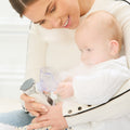A woman helps a baby use Dr. Talbot's Portable Ultrasonic Mesh Nebulizer indoors, holding the device as the baby places the mask on their face. Both are in light-colored clothes.
