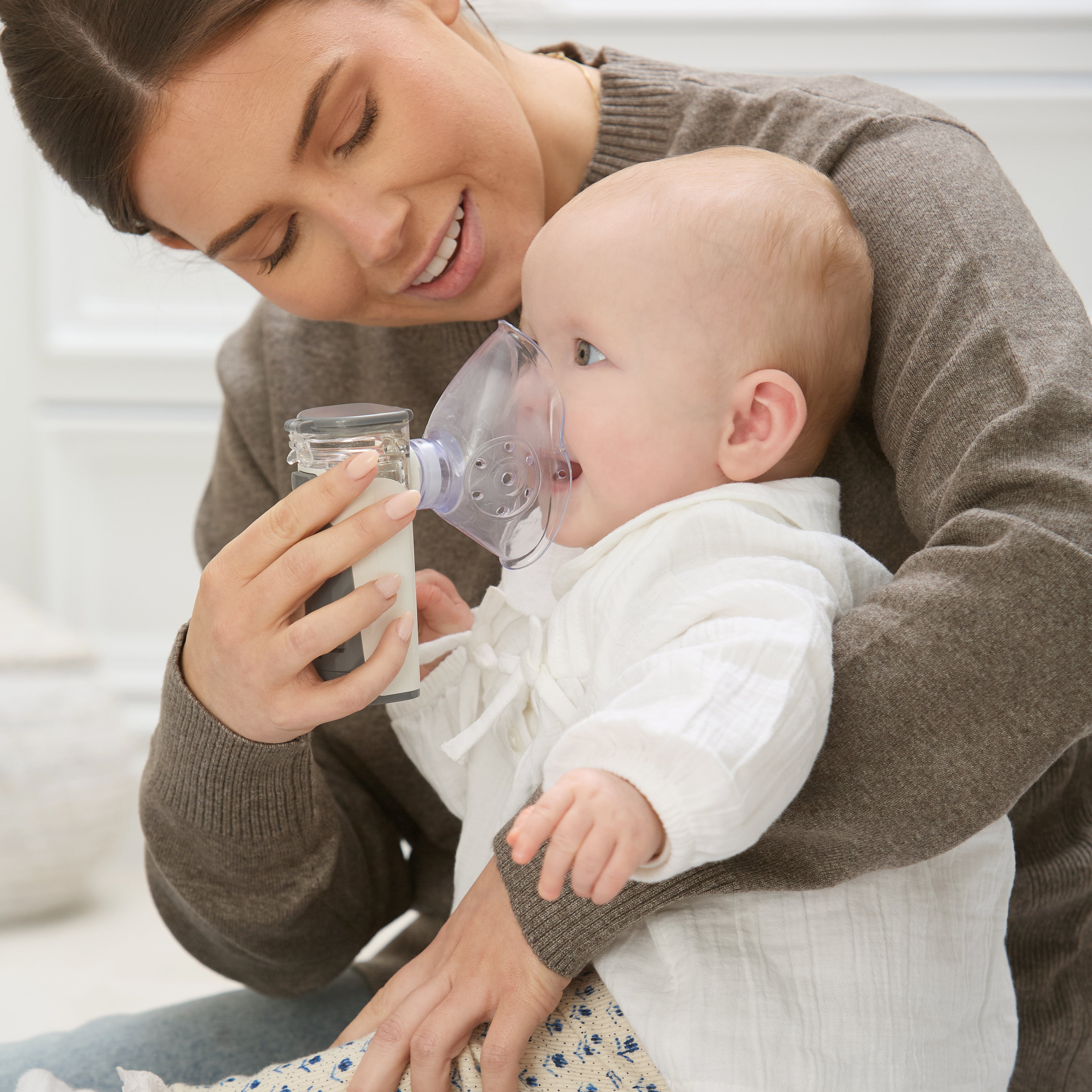 A woman assists her baby, dressed in white, while using Dr. Talbot's Portable Ultrasonic Mesh Nebulizer for respiratory relief. The baby sits calmly on her lap as she smiles and looks at the child.