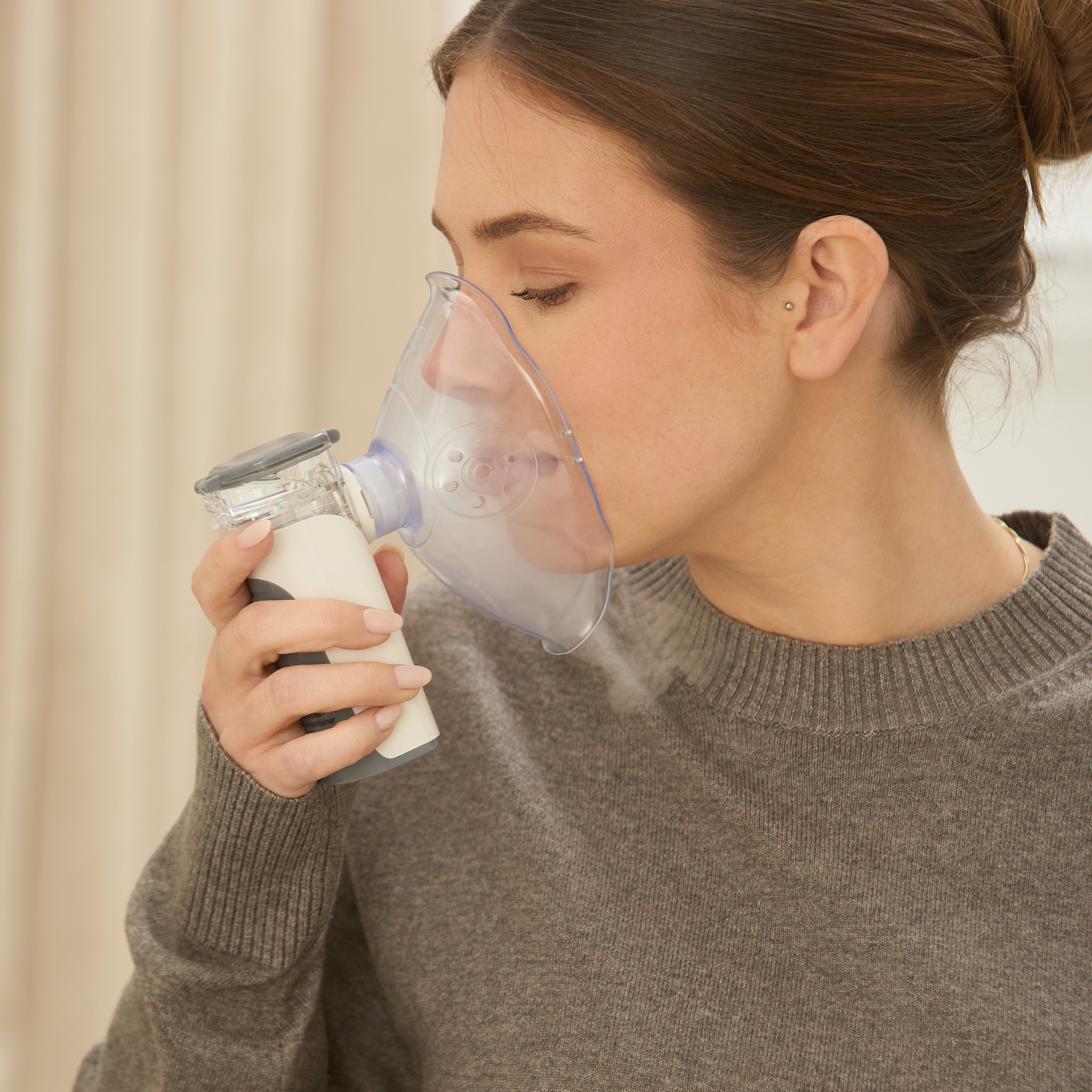 A woman in a gray sweater uses Dr. Talbot's Portable Ultrasonic Mesh Nebulizer with a clear face mask, inhaling medication for respiratory relief.