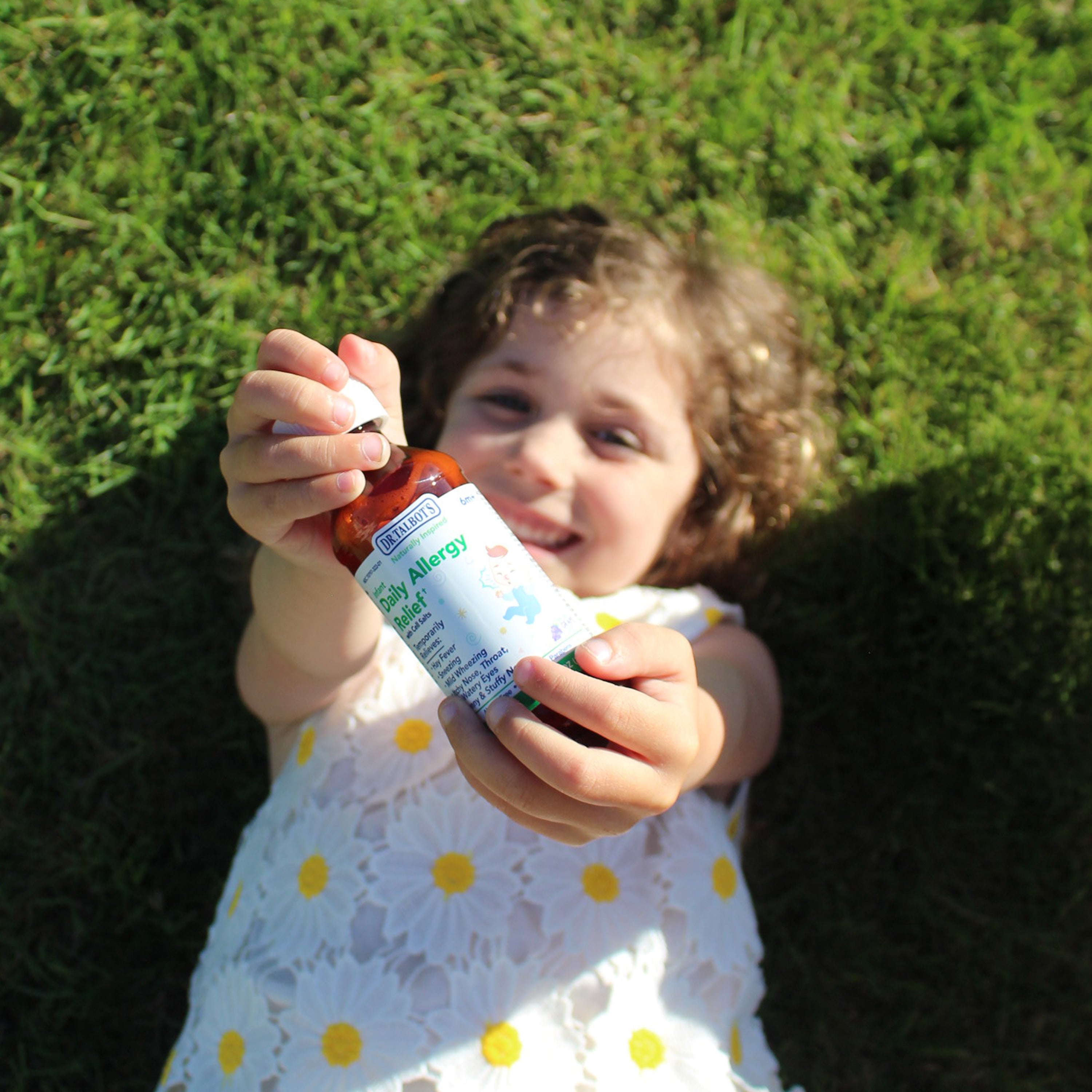 A young child lying on the grass holds a bottle of Dr. Talbot’s Infant Daily Allergy Relief toward the camera.