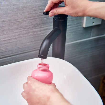 A person fills Dr. Talbot's Mom Postpartum Peri-Bottle with water from a modern black faucet over a white sink in a bathroom with gray tiled walls.