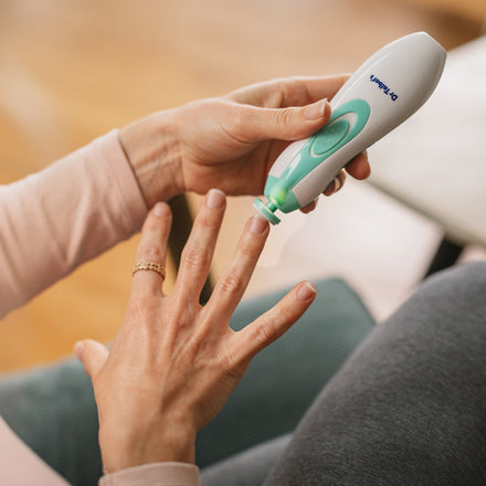 A person seated on a blue cushion uses Dr. Talbot's Baby Nail Trimmer with Hygienic Case on their fingernail while wearing a pink long-sleeve top and a gold ring. The white and green device is part of the baby manicure set.