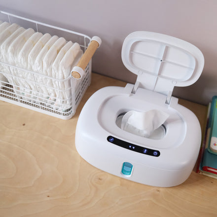 A Dr. Talbot's Touch-Free Wipe Warmer with its lid open is placed on a wooden surface beside a wire basket of diapers and a stack of books.