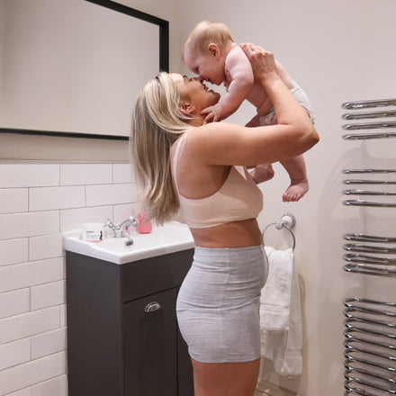 A woman in a beige sports bra and gray shorts lifts her smiling baby in a bathroom. Dr. Talbot's Mom Disposable Postpartum Panties (8 Pack) are visible near the sink and towel rack as they share a joyful moment.