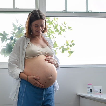 A pregnant woman stands by a window with green leaves outside, smiling and touching her bare belly. She wears a beige bra, blue pants, and an open white shirt. Dr. Talbot's Pregnancy Stretch Mark Cream is on the counter nearby.