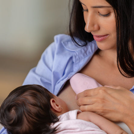 A woman in a blue shirt breastfeeds her dark-haired baby, wrapped in a light blanket, while using Dr. Talbot’s Mom Lactation Massager (pink) on her breast to help prevent clogged milk ducts.