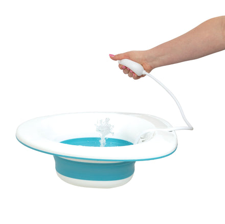 A hand uses Dr. Talbot's Mom Postpartum Sitz Bath, spraying water from a white nozzle into a blue and white plastic basin for perineal cleansing, against a white background.
