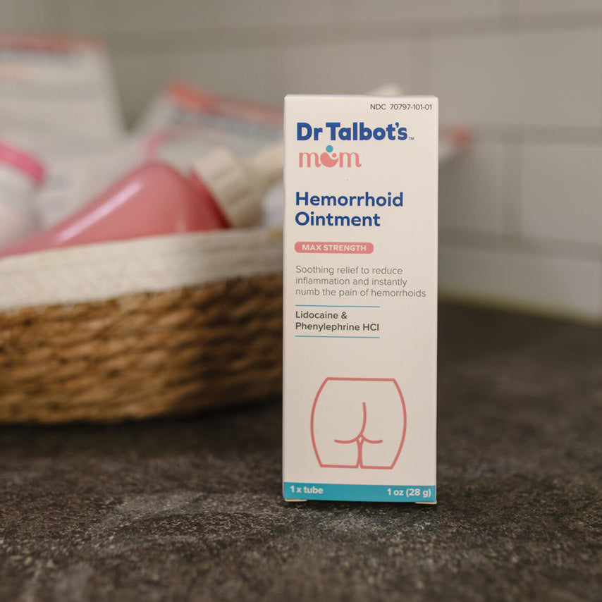 A box of Dr. Talbot's Mom Pregnancy & Postpartum Hemorrhoid Ointment sits on a countertop, its packaging featuring an outline drawing of buttocks and highlighting hemorrhoid relief for pregnancy, with baby bottles blurred in the background.