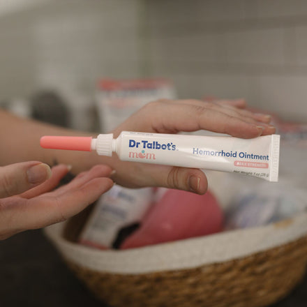 A hand holds a tube of Dr. Talbot’s Mom Pregnancy & Postpartum Hemorrhoid Ointment with a pink applicator in a kitchen or bathroom, and a woven basket is visible in the blurred background.