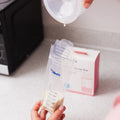 A person pours breast milk into a clear, puncture-resistant MilkFresh Breastmilk Storage Bag by Dr. Talbot’s Mom on a kitchen counter, with a box of storage bags and a bottle warmer in the background.