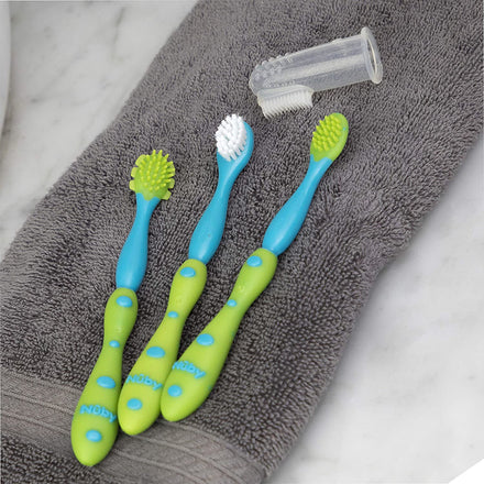 Dr. Talbot's Oral Care Set 4-Stage System, featuring three colorful baby toothbrushes and a clear fingertip toddler brush with green and blue circular grip handles, is displayed on a folded gray towel atop a marble surface.